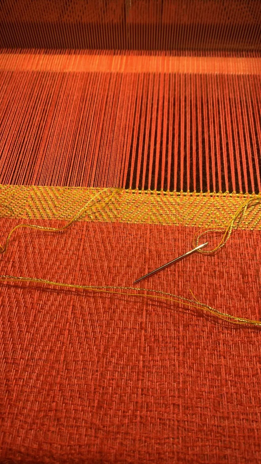 Gros plan sur un tissage rouge brique à chevrons dorés, une aiguille dorée piquée dans l'ouvrage